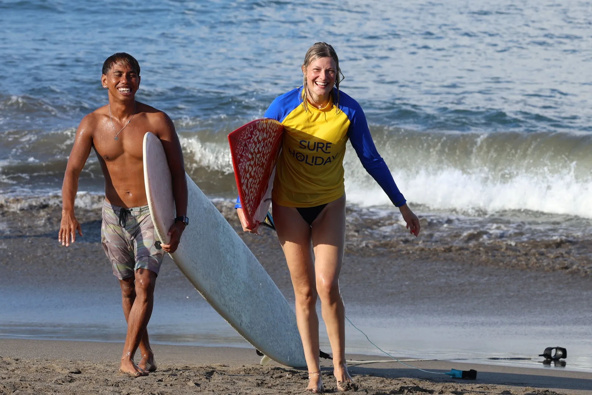 A Surf Holiday Bali coach and student walking up the beach with their boards, all smiles