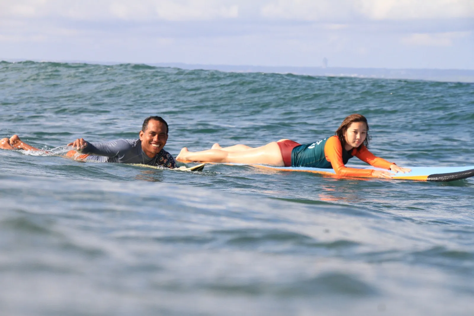A coach and student paddling out side by side on their soft-top boards, both smiling