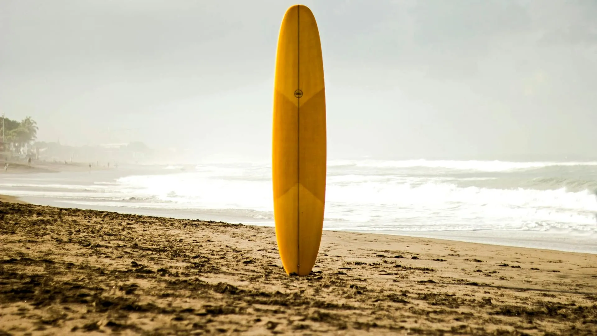 A yellow longboard standing upright in the sand on Batu Bolong beach, Canggu, with waves rolling in behind
